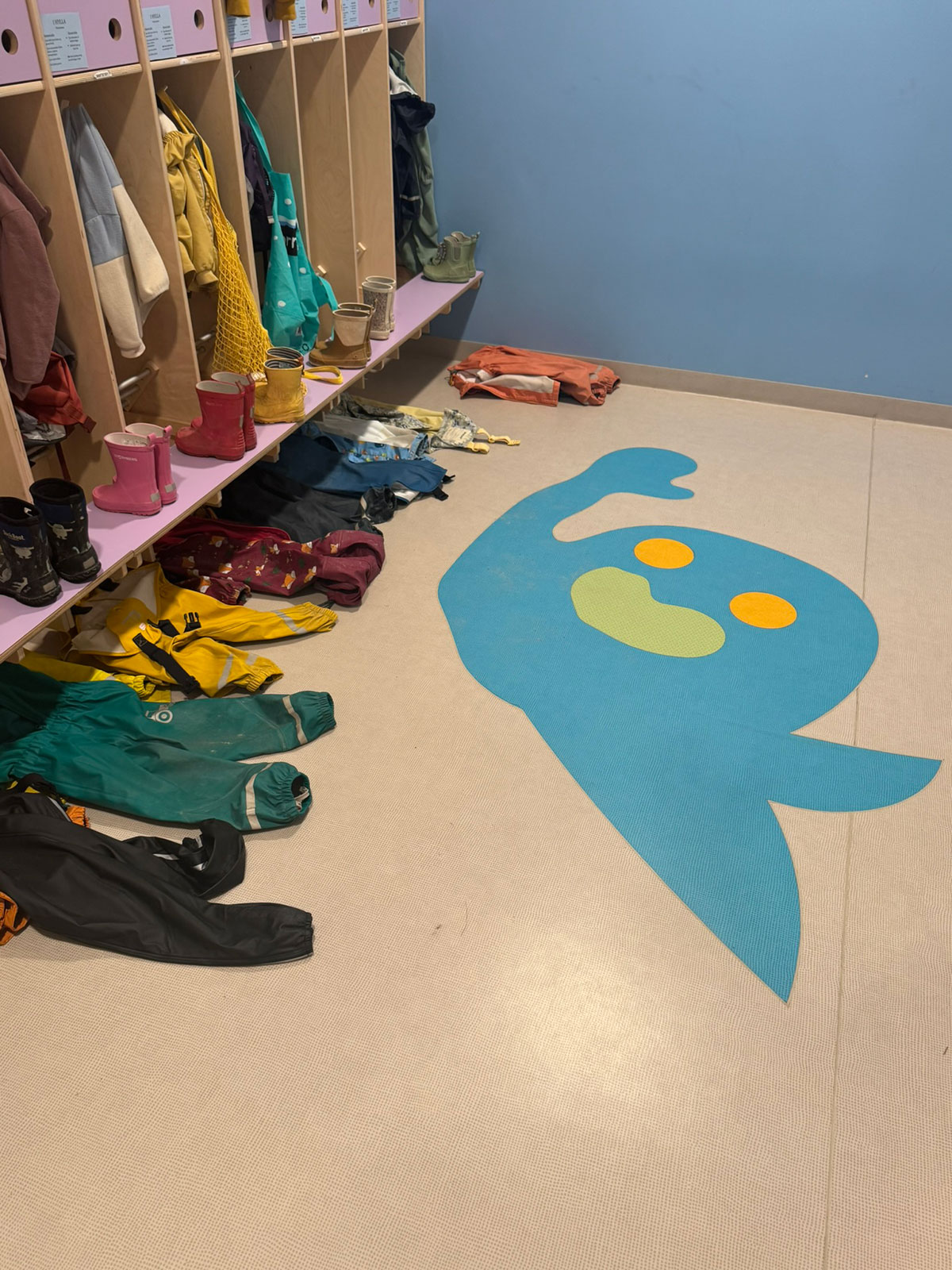 en blå spøkelsesfigur midt på gulvet i garderoben eller gangen i en barnehage. Til venstre er det åpne garderobeskap med klær hengende og strødd utover gulvet.