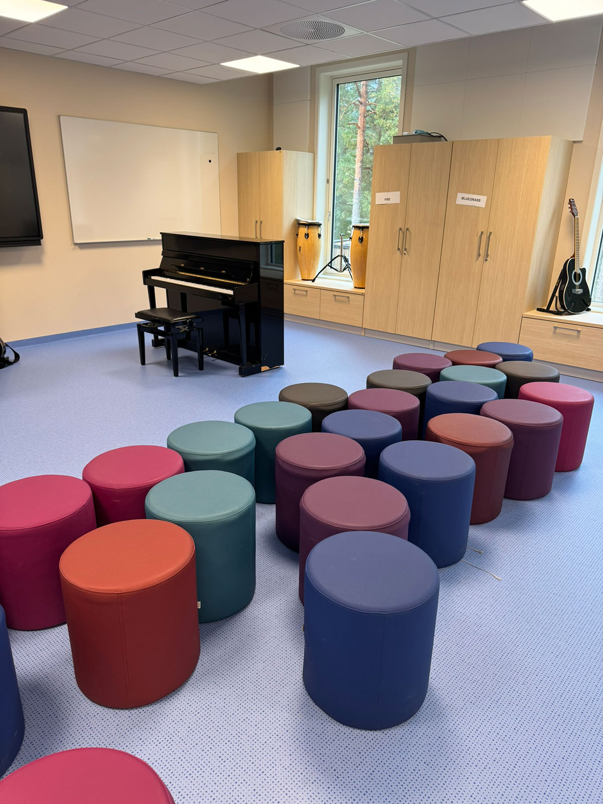 Et musikkrom med et sort piano i bakgrunnen og sylinder formet puffer i mange ulike former rett på det blå gulvet