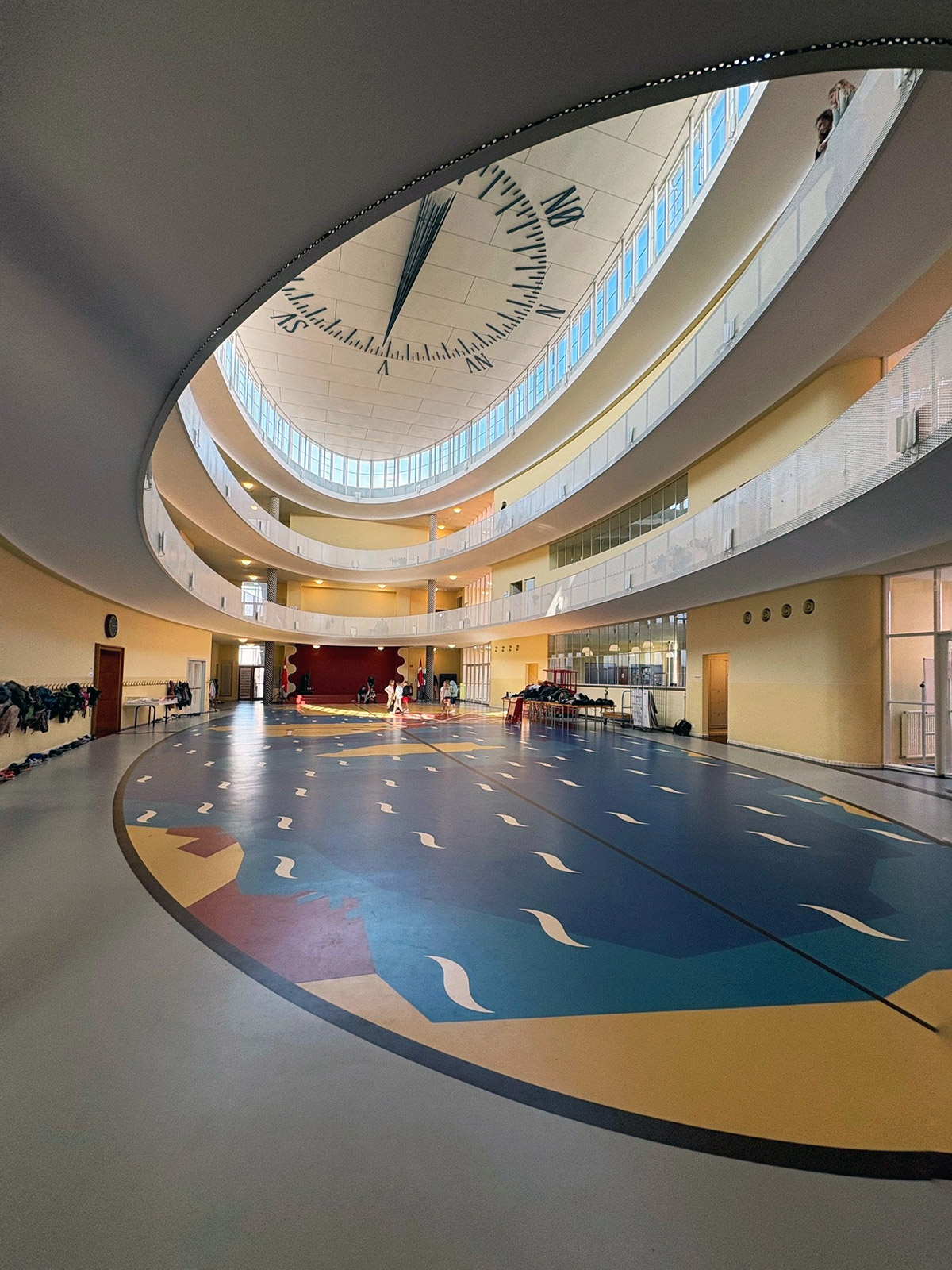 Interiørbilde av en stor, lys skolebygning med et ovalt atrium og flere etasjer med åpne gallerier. I taket er det et stort ur, og gulvet har et fargerikt mønster i blått, gult og rødt. Noen personer står i bakgrunnen i det åpne rommet.