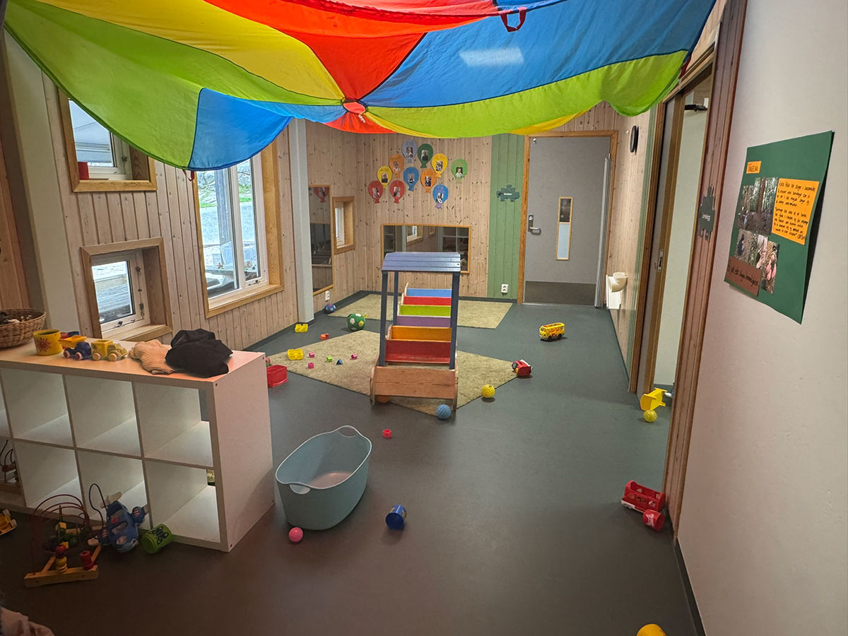 Lekerom i en barnehage med hyller og skap, leker strødd utover gulvet og et fargerikt seil festet i taket