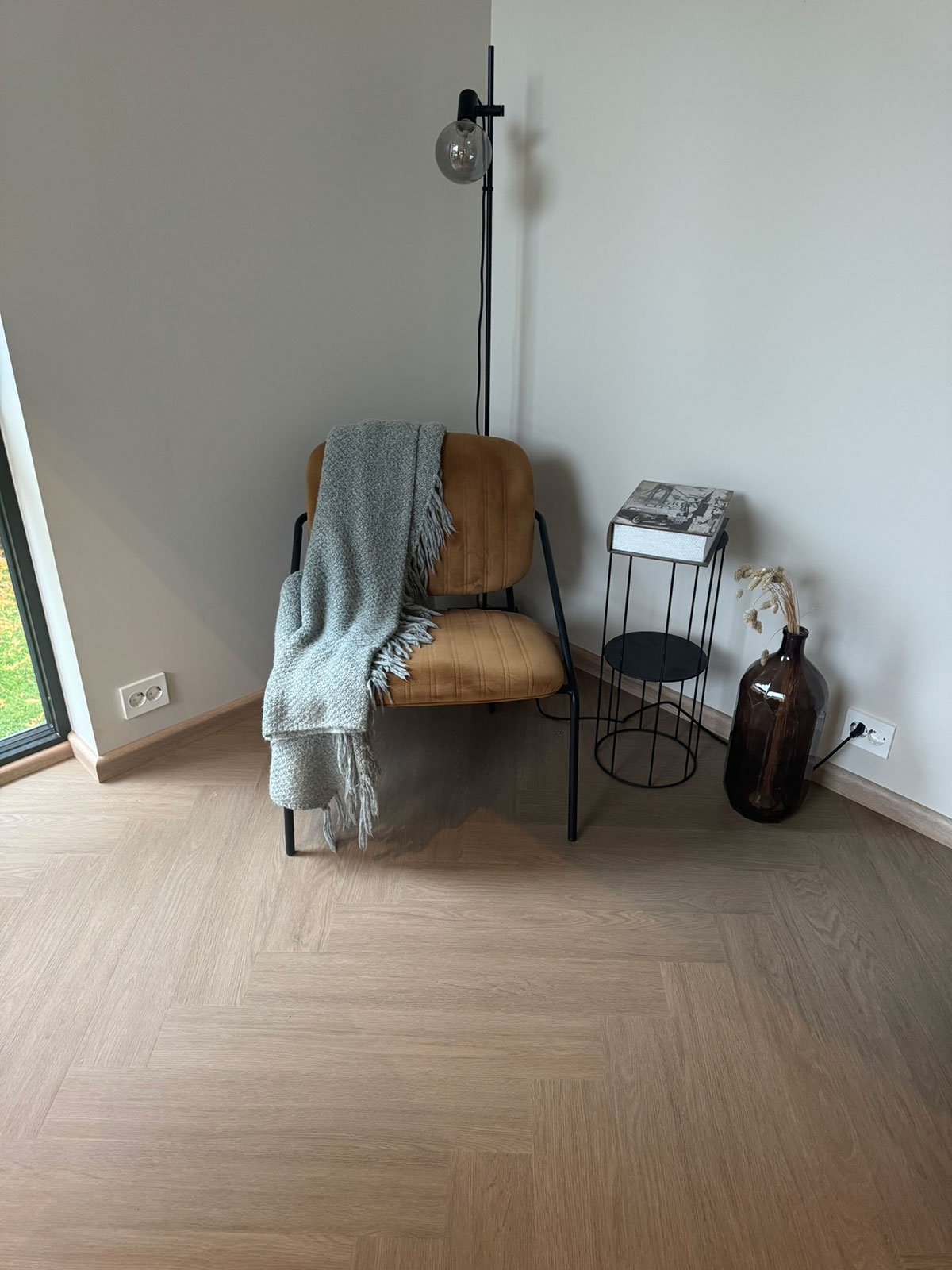 LVT-gulv i lys eikefarge lagt i fiskebensmønster, vist i et moderne hjørne med stol, pledd, gulvlampe og dekorative detaljer i varme toner.