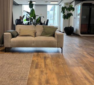 Moderne kontorlokale med beige sofa, grønne planter, trestrukturert gulv og åpent arbeidsområde i bakgrunnen.