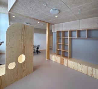 Lyst oppholdsrom med grått gulvbelegg, kryssfinerinnredning, buet lekeskillevegg og åpne trehyller.