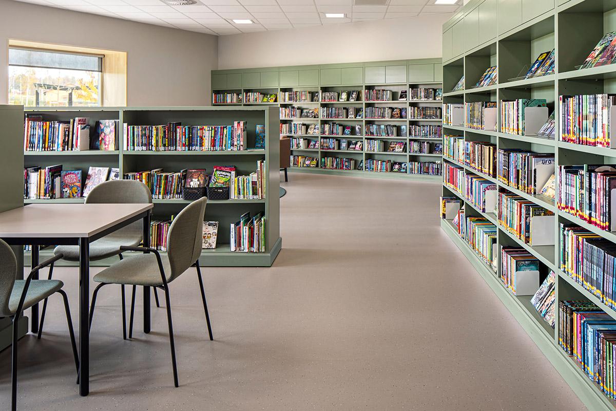 bibliotek på en skole med bokhyller i grønt til høyre og bord og stoler til venstre