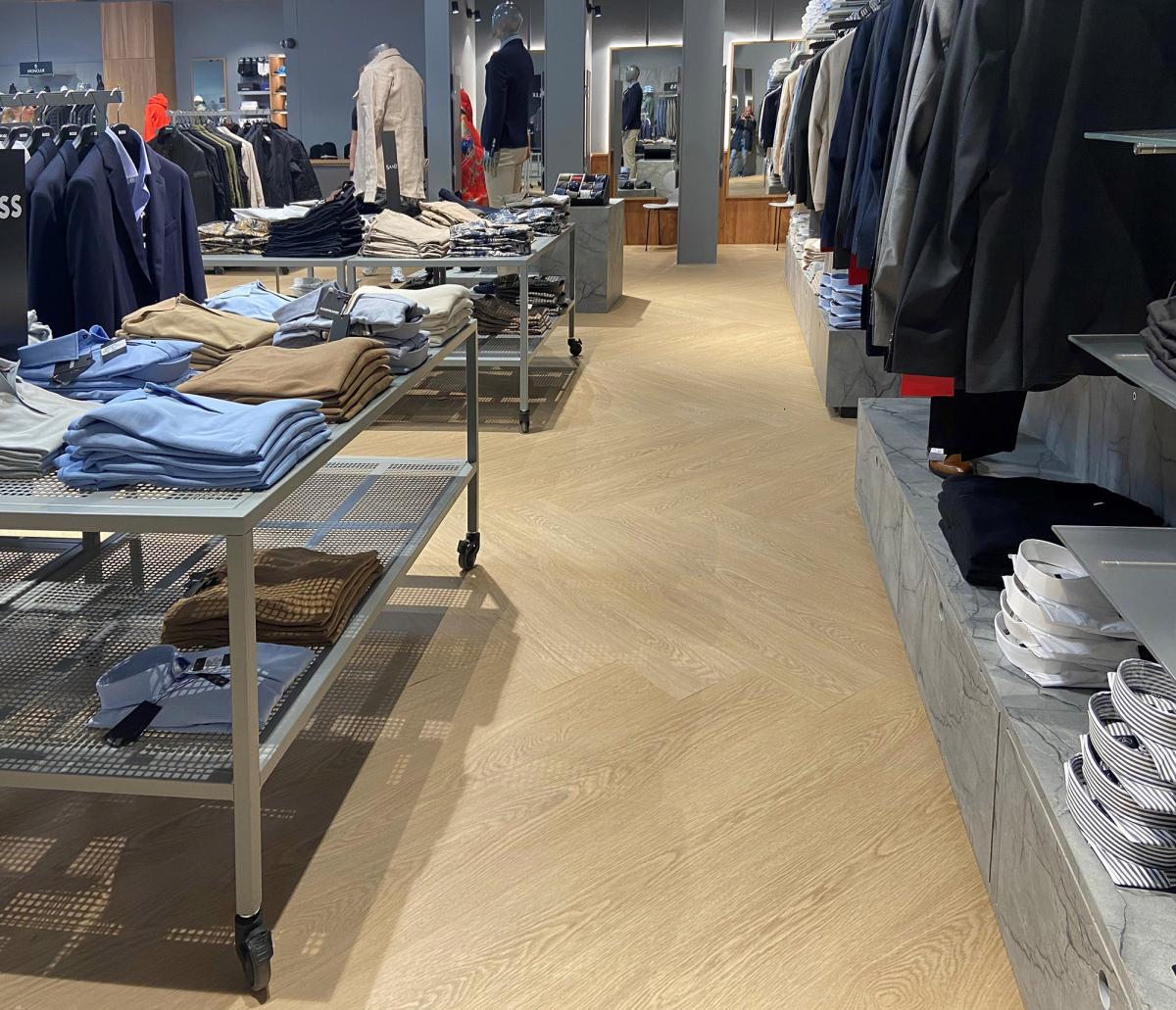 flere bord med klær til venstre i en klesbutikk. Hyller med klær til høyre. Lyst eike-lignende gulv
