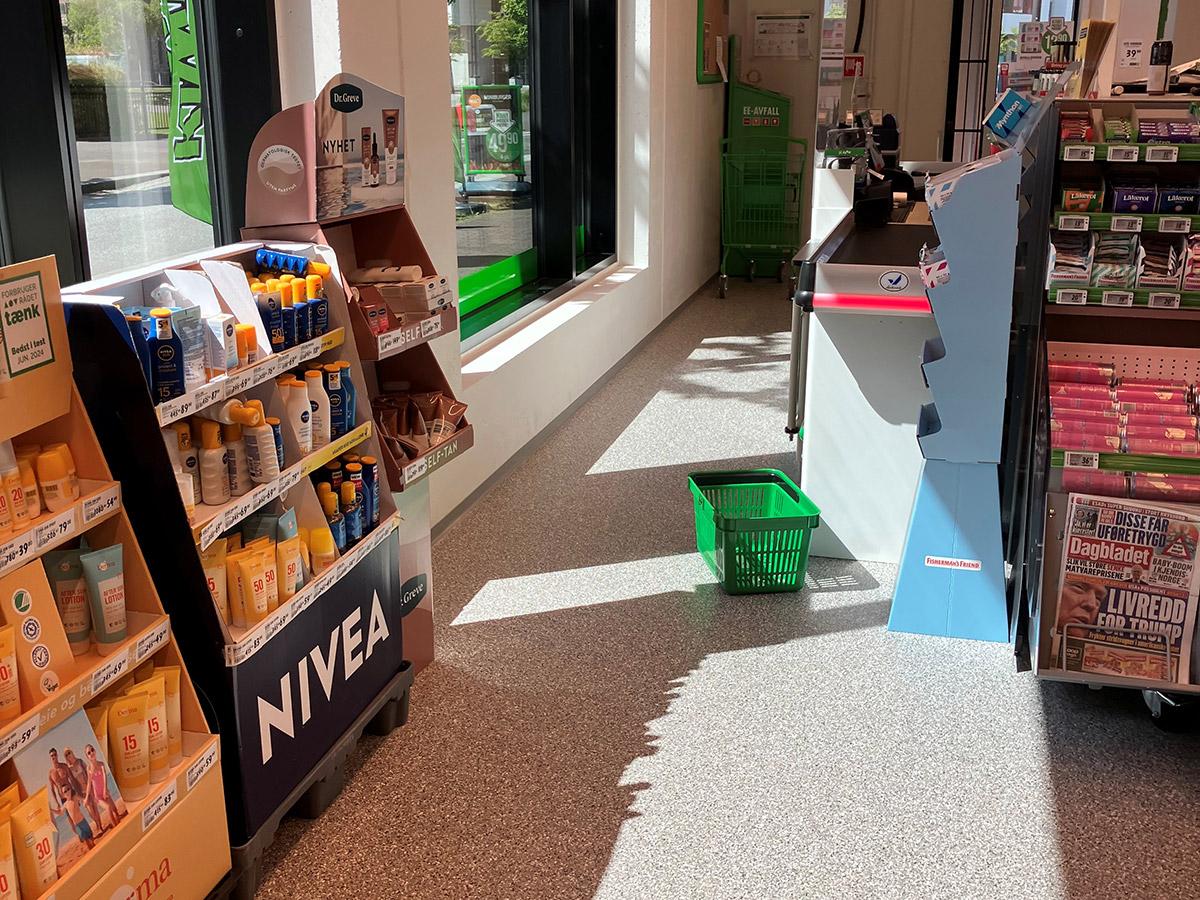 kasseområde i en kiwi butikk med hyller til venstre og en grønn handlekurv stående på gulvet