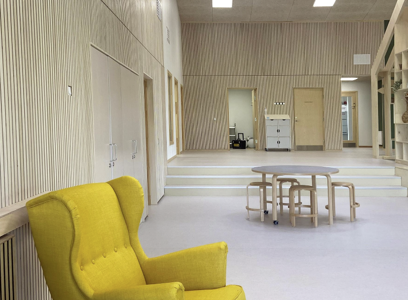 fellesområdet i en barnehage med trevegger og lyst gulv. I forgrunnen står det en gul lenestol i midten en sittegruppe med bord og stoler