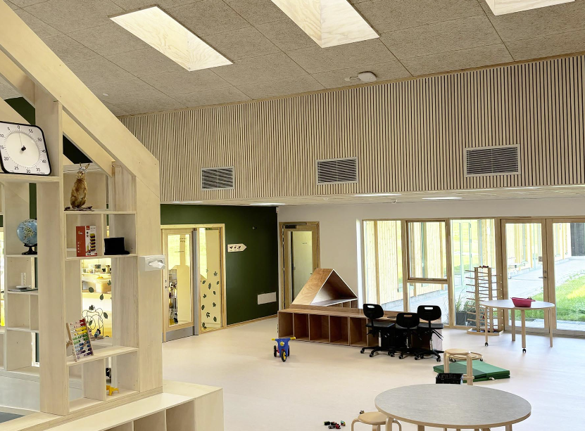 lekerom i en barnehage med god takhøyde. Til venstre ser man et lekerom bygget som et hus, i forgrunnen et bord og i bakgrunnen vinduer fra gulv til tak med utsikt ut til gressplenen