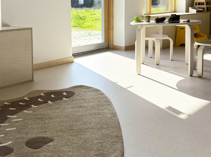 nærbilde av gulvet i et rom i barnehagen. Et brunt teppe ligger til venstre i bildet og til høyre ser man bord og stoler