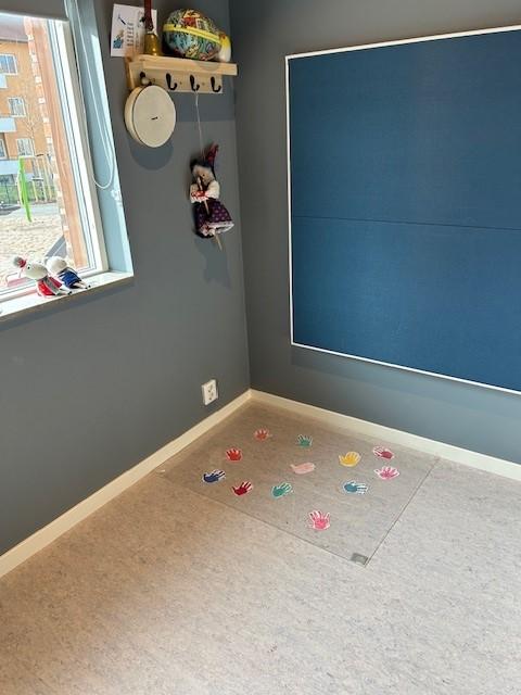 hjørnet i et barnerom i en barnehage med grå vegger og en blå oppslagstavle i tekstil som dekker nesten hele den ene veggen