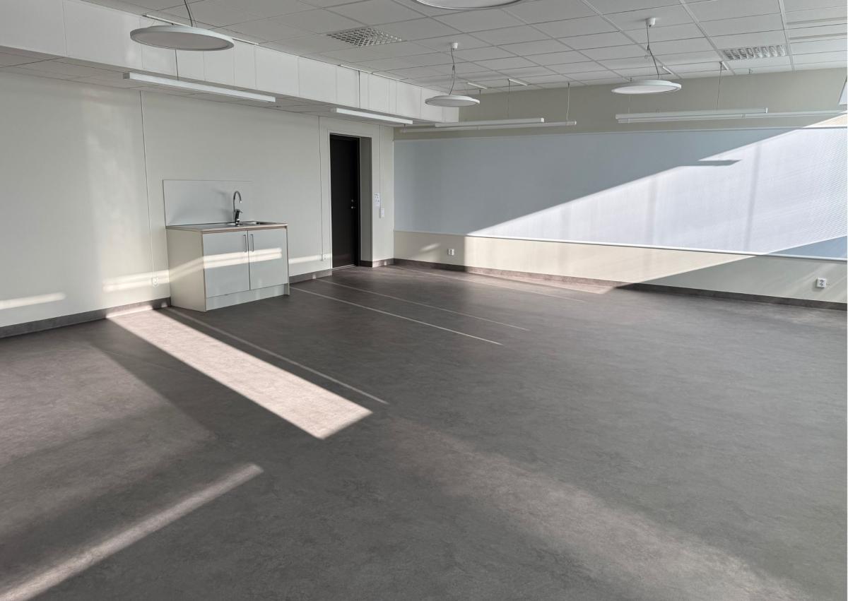 et tomt klasserom med lyse vegger, et grått gulv, en lang tavle til høyre i bildet og en kort kjøkkenbenk på veggen til venstre ved siden av inngangdøren 