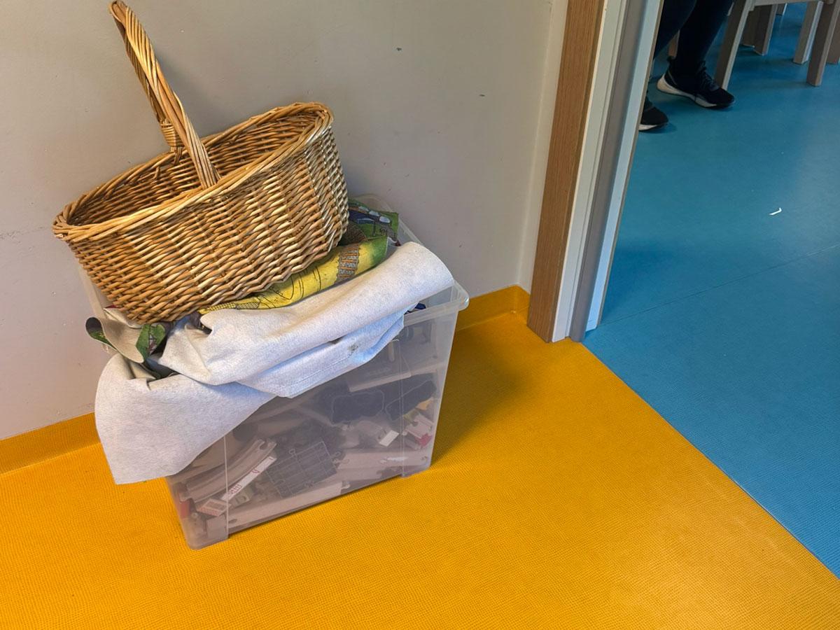en plastkasse og en kurs står inntill veggen på et gult gulv. til høyre kan man se at neste rom har blått gulb