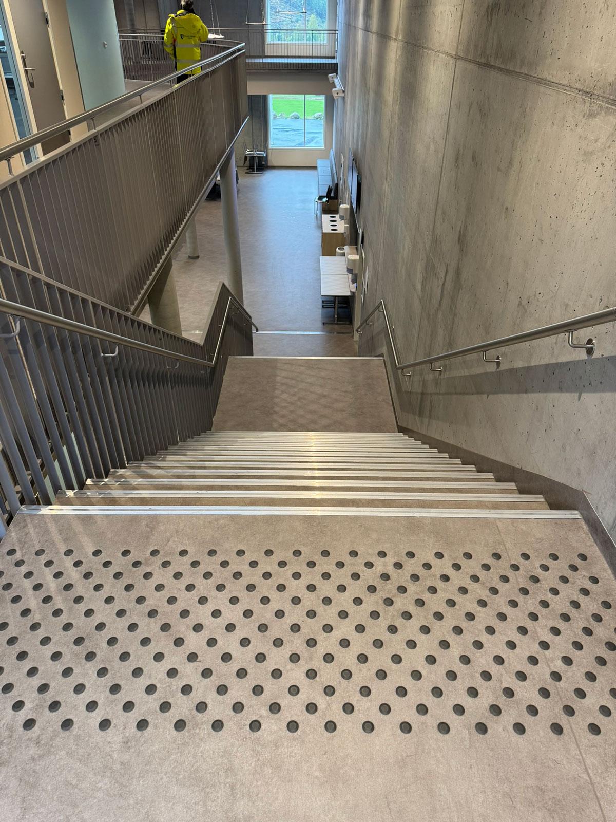 En trapp nedover til etasjen under. I forgrunnen ser man skilhemmende sorte runde dotter i gulvet. 