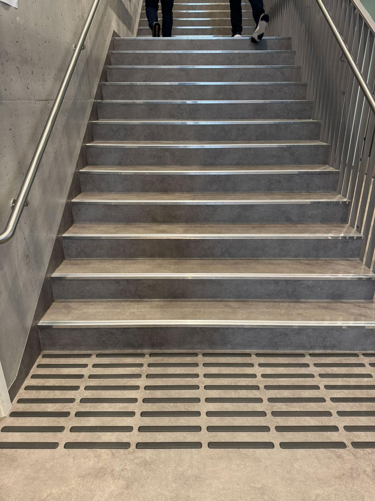 En trapp opp til etasjen over. I forgrunnen ser man avlange sorte skilhemmende opphøyninger i gulvet. Øverst i trappen kan man så vidt se benene til to personer som går oppover