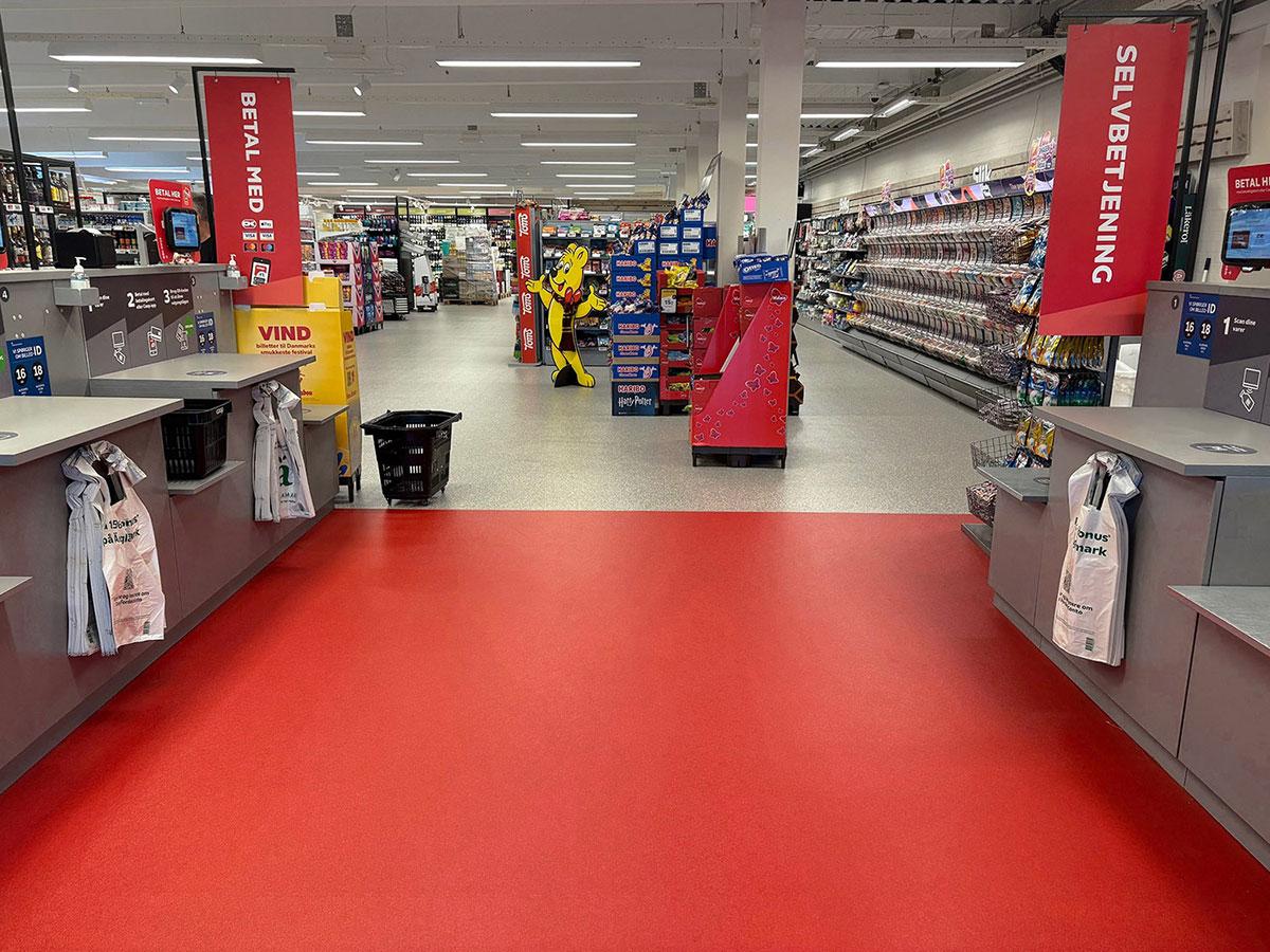 selvbetjente kasser i en matvarebutikk med kasser både til høyre og venstre. Gulvet rundt kassene er rødt og markerer området. Gulvet ellers i butikken er grått