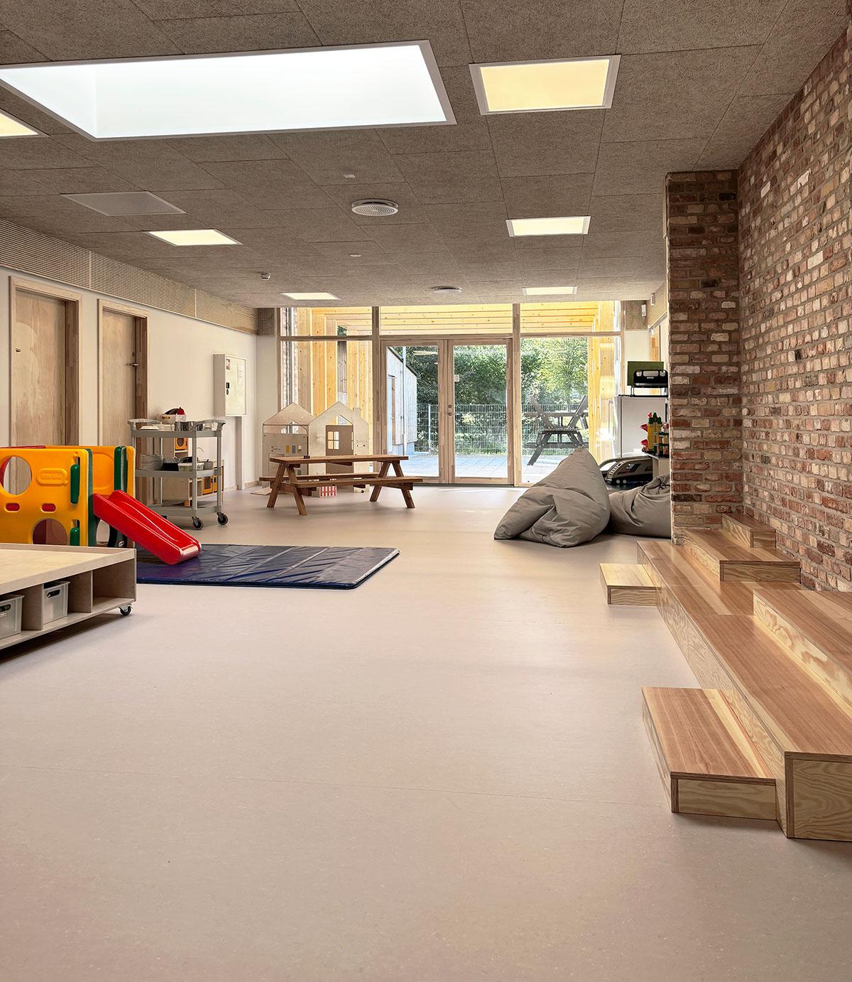 oppholdsrom på et barnehjem med klatrestativ, benk og lekehus til venstre. Sakkosekk og trappetrinn foran en mursteinsvegg til høyre. Lyst og beige gulv