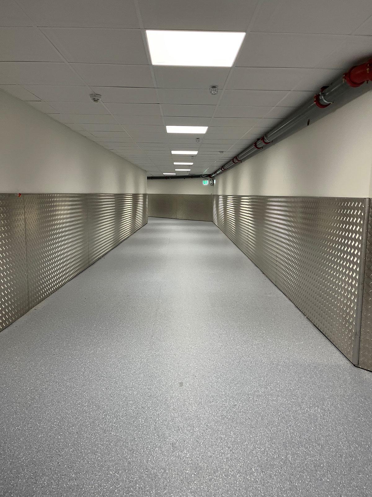 en lang gang på et sykehus med grått gulv og beskyttelsesplater på veggene