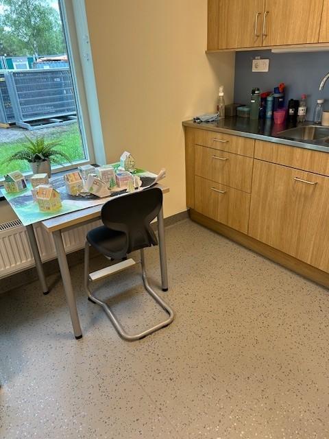 kjøkkenet på en skole med kjøkkenbenk til høyre og en stol stående inntil et bord til venstre. på bordet står det mange papirhus