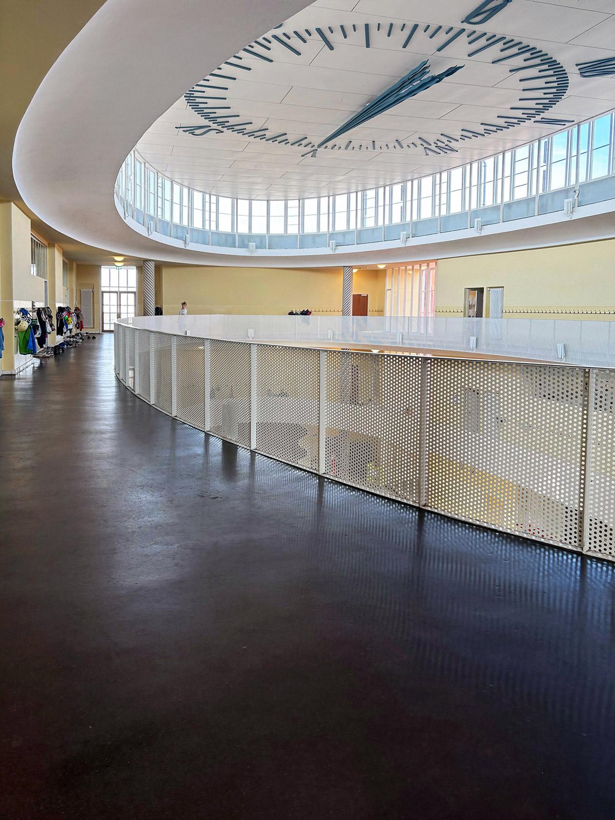 Lys og moderne skolebygning med buet korridor og perforert rekkverk i metall. Over atriet sees et stort takur med markerte visere og tall, omgitt av vinduer som slipper inn naturlig lys. En person står ved rekkverket, og garderober med klær ses langs veggen.