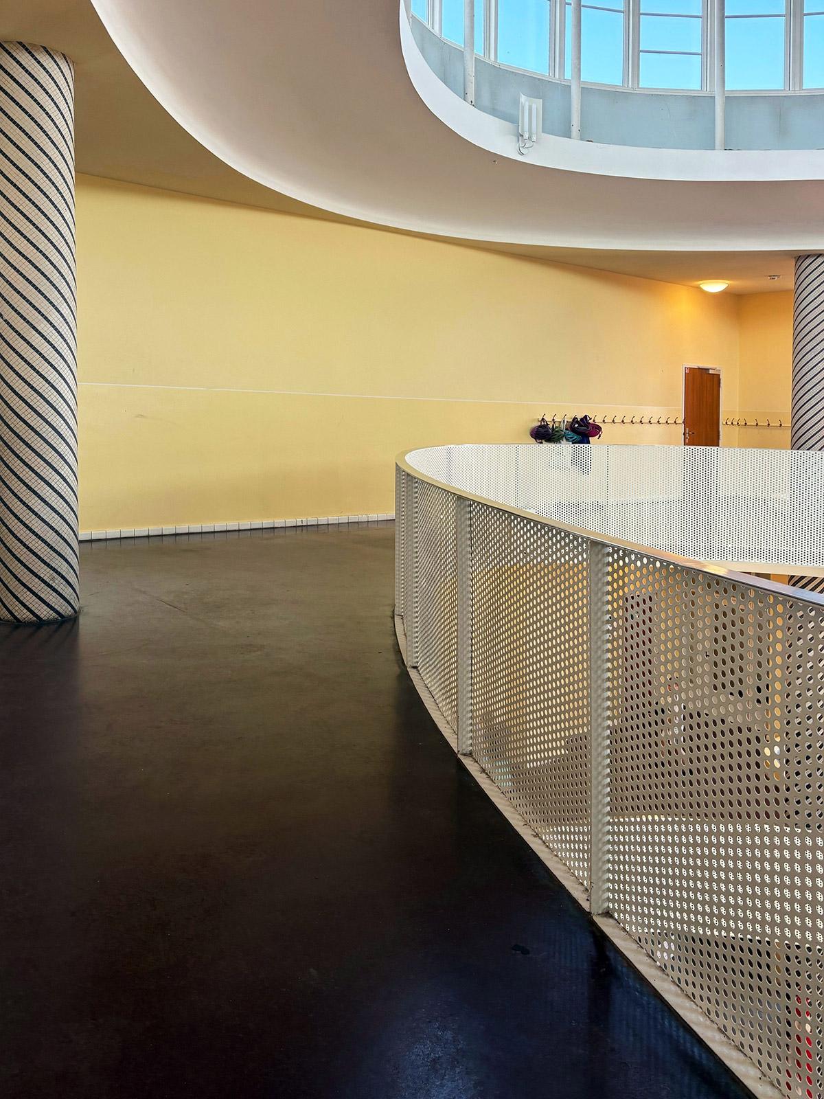 Detaljbilde fra en moderne skolebygning med buet gangbro og perforert metallrekkverk. Veggene er malt i lysegult, og et stort vindusparti slipper inn naturlig lys ovenfra. En søyle med diagonale striper står til venstre i bildet.