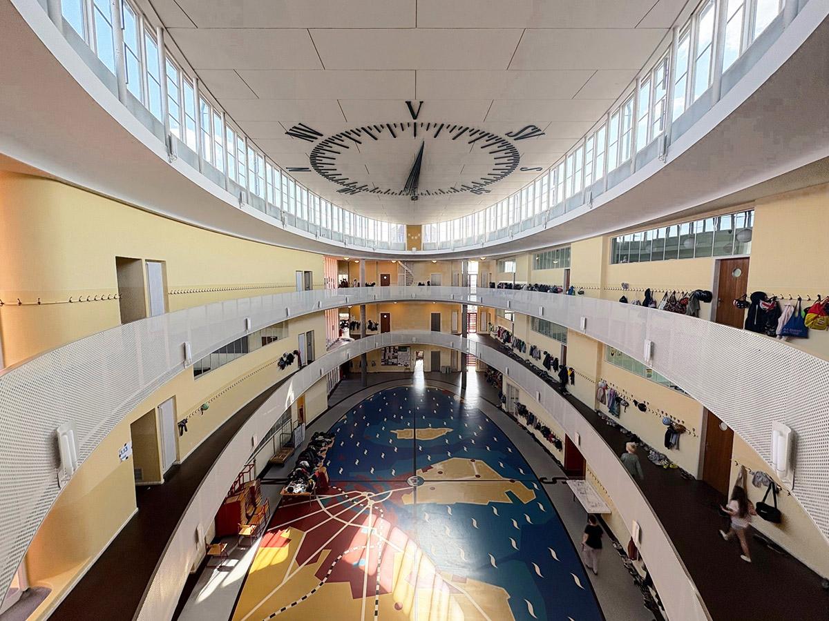 Oversiktsbilde av et lyst og åpent atrium i en moderne skolebygning. Midten av rommet har et fargerikt gulvmønster i blått, gult og rødt, mens de buede gangene i flere etasjer omkranser atriet. I taket ses et stort ur med markerte tall og visere. Elever og klær henger langs rekkverkene.