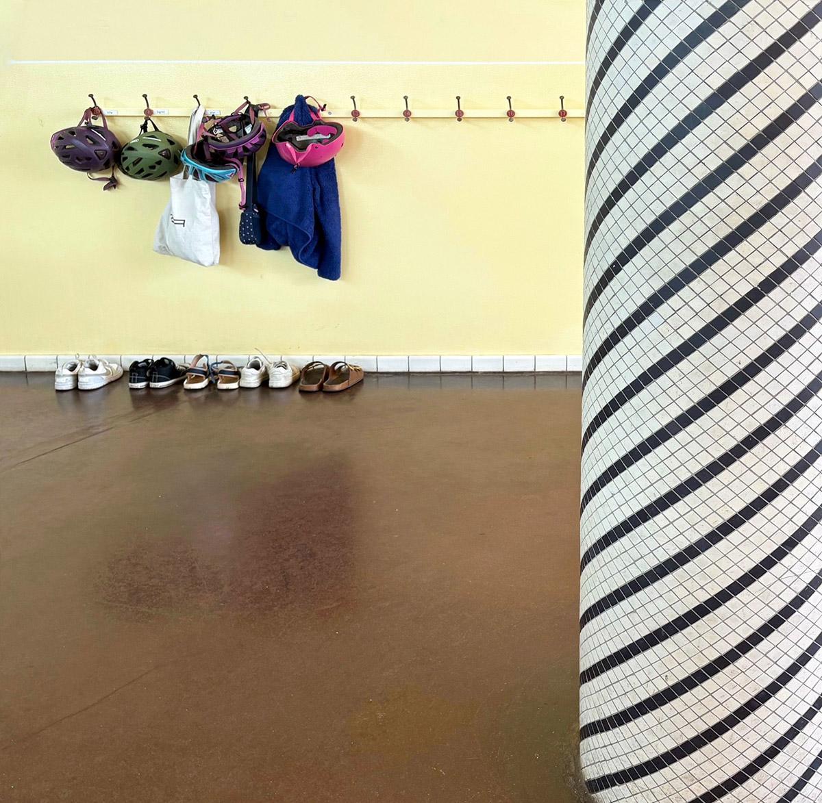Nærbilde fra en skolekorridor med mørkt linoleumsgulv og lysegul vegg. Langs veggen henger hjelmer, klær og vesker på knagger, og sko står pent oppstilt på gulvet. Til høyre i bildet sees en søyle med diagonalt stripemønster i mosaikk.