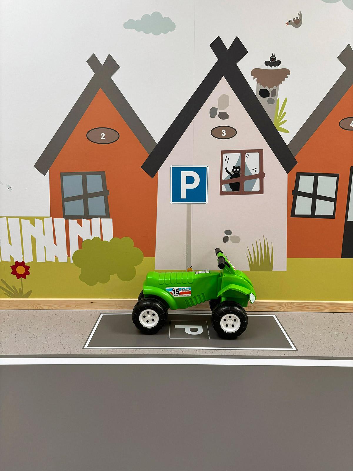 en grønn lekebil stående parkert til venstre foran en lang vegg. På veggen er det dekorert med tegninger av ulike hus