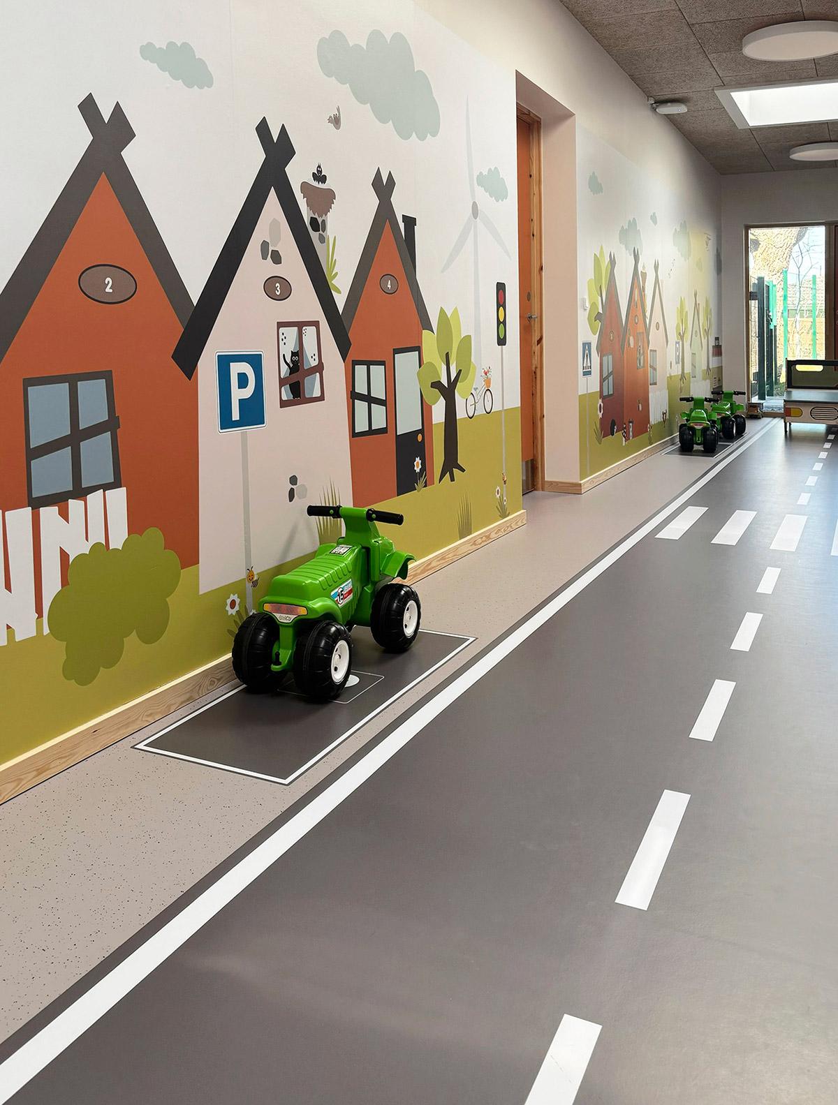 en grønn lekebil stående parkert til venstre foran en lang vegg. På veggen er det dekorert med tegninger av ulike hus. På gulvet er det dekorert med imitasjon av vei delt i to veibaner