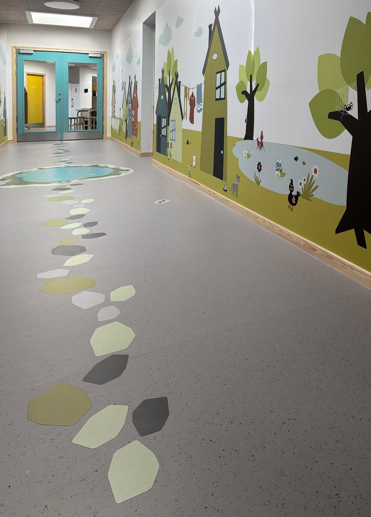 en gang i en barnehage hvor veggen til venstre er dekorert med tegninger av hus og trær. På gulvet er det grønne felt formet som steiner som danner en sti på gulvet
