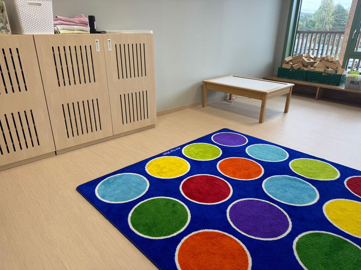 Et blått teppe med masse sirkler i ulike farger liggende på et gulv i en barnehage. Et skap til venstre og vindu til høyre