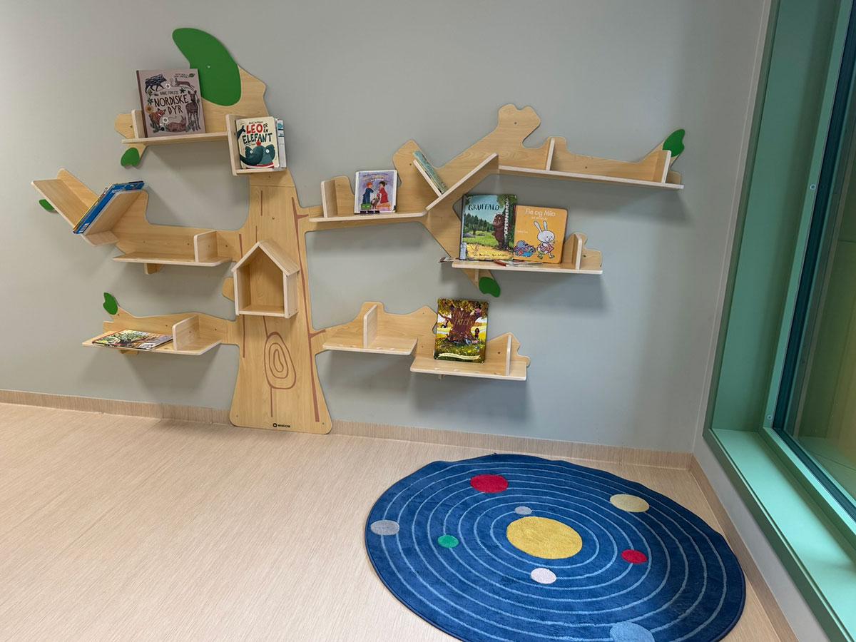 En hylle i treverk formet som et tre med plass til bøker og leker på greinene som streker seg både til høyre og venstre. På gulvet ligger det et rundt, blått teppe med fargede sirkler