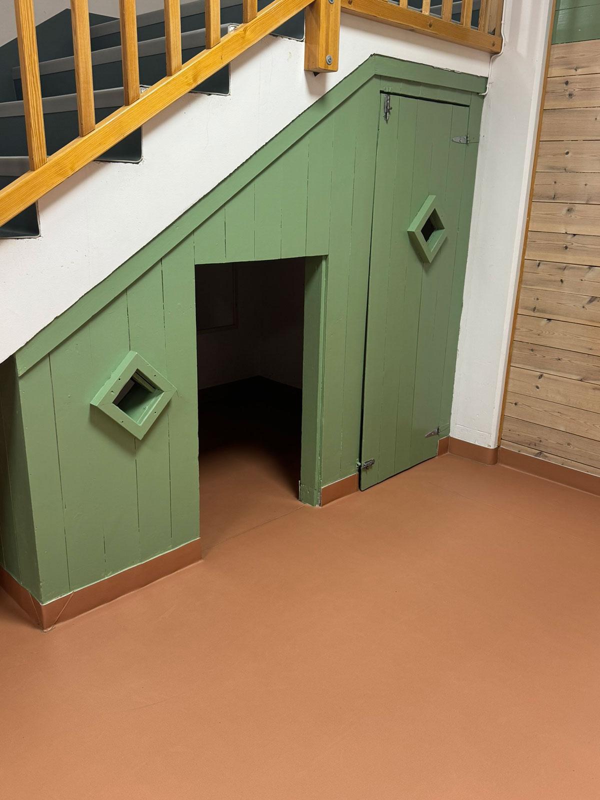 Et lekerom med grønne vegger, liten dør og vindu bygget inn under en trapp i et hjørne