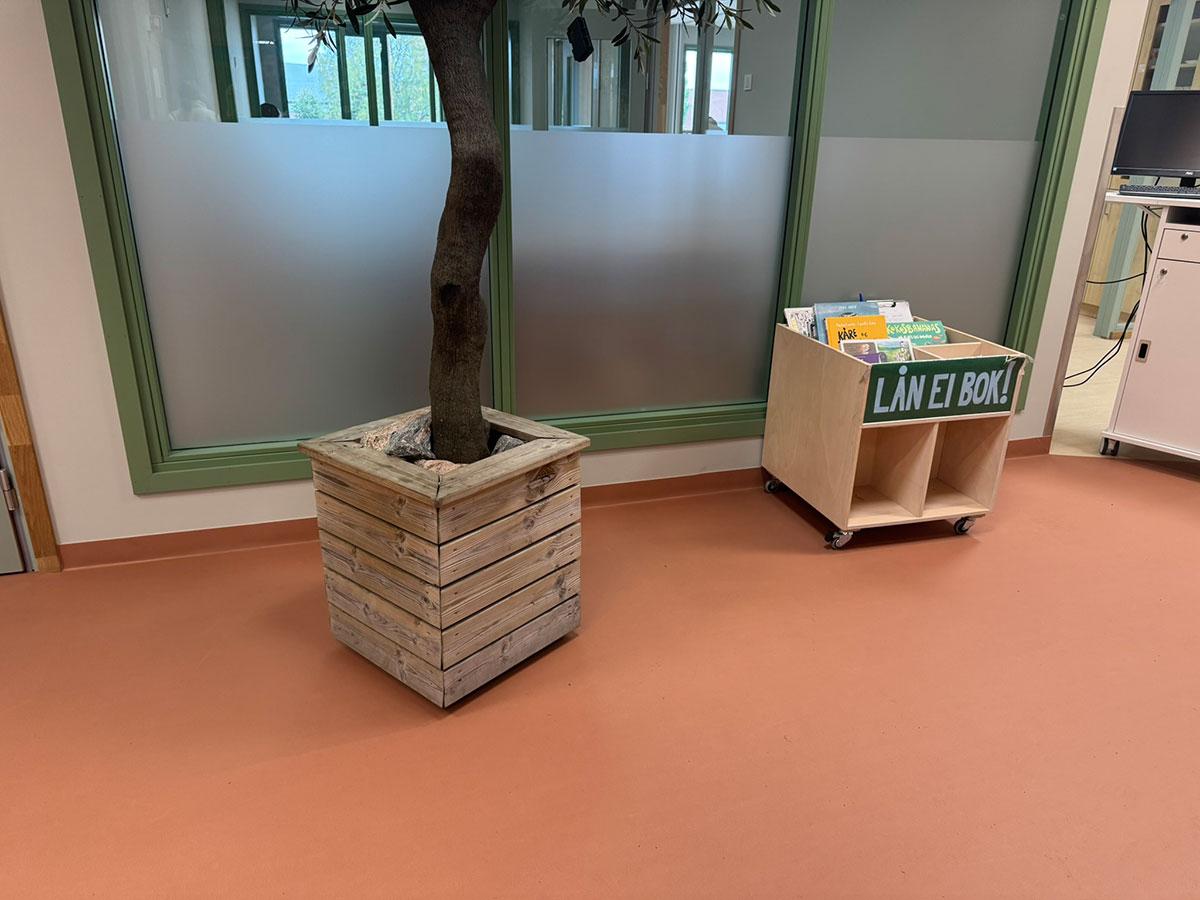 fellesgang i en barnehage med en kvadratisk blomsterkasse med et tre stående i. Til høyre ser man et lite skap på hjul med bøker