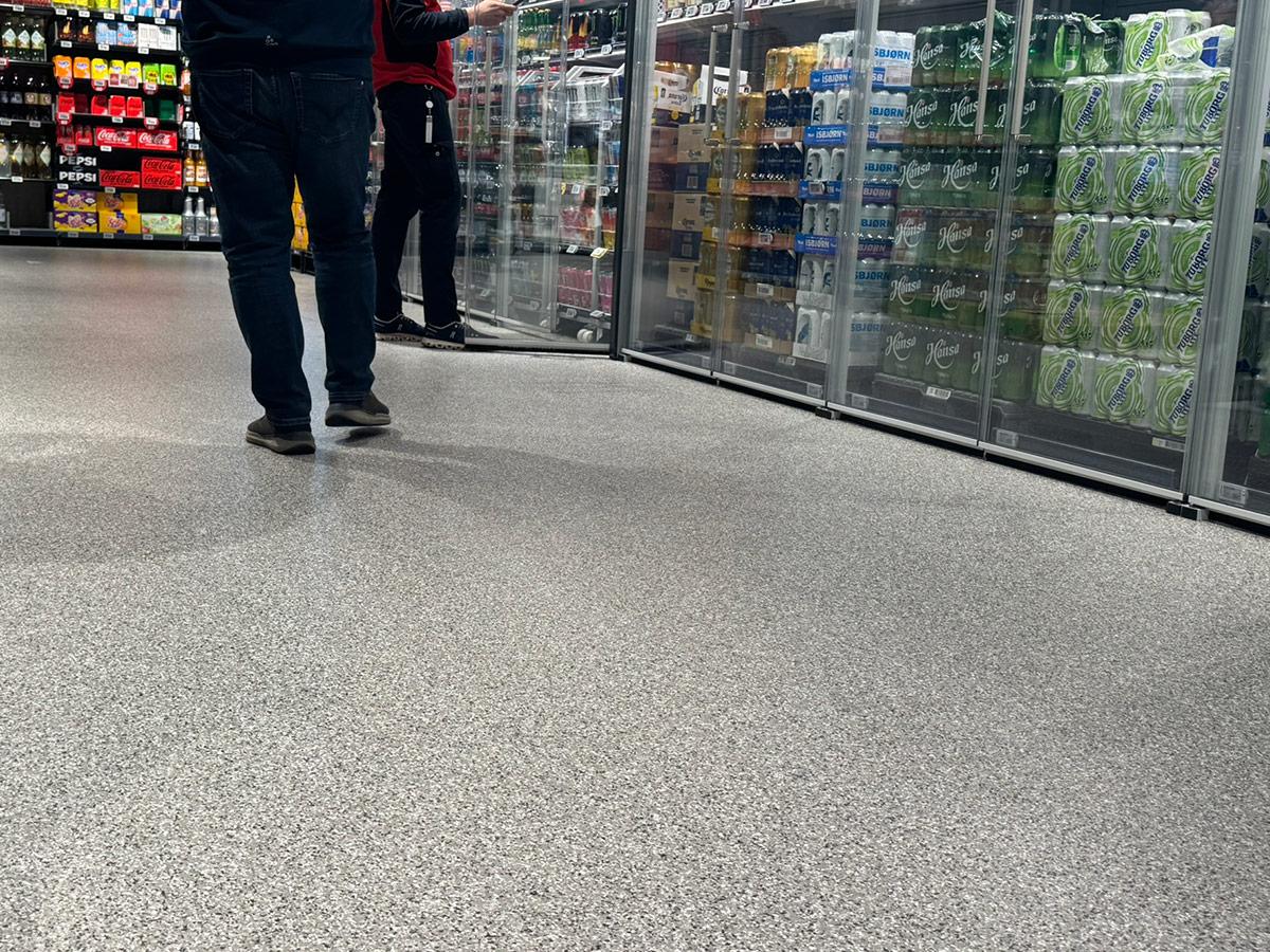 butikkgulv nedenfra foran kjøleskap med drikke. To mennesker går på guvlet hvor man kun ser fra livet og ned