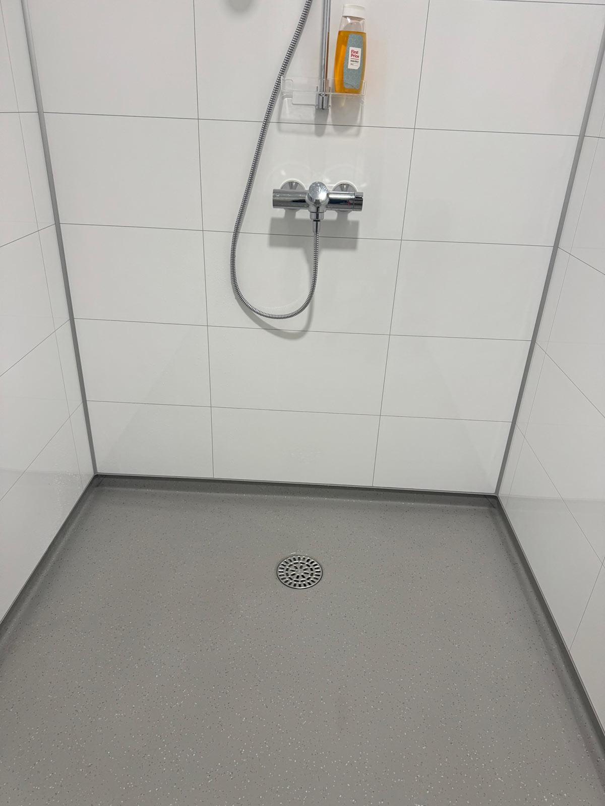 en dusj med hvite fliser, grått gulv og sluk i midten