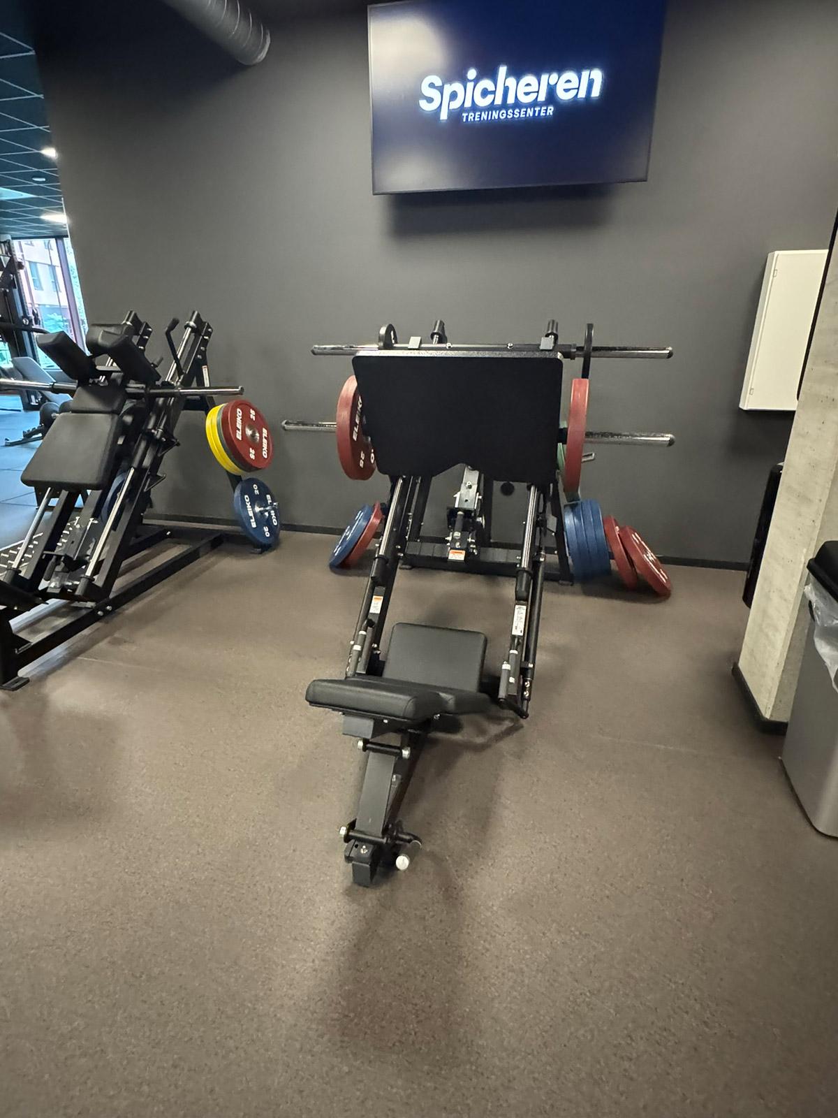 Treningsapparat for beinpress foran vegg med Spicheren-logo på treningssenteret, med robust gulvbelegg tilpasset tung bruk.
