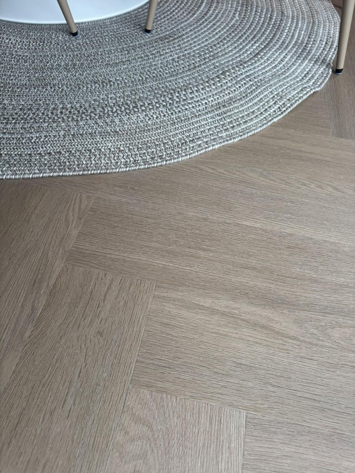 Lyst LVT-gulv i fiskebensmønster, vist sammen med et rundt, vevd teppe og lyse stolben som fremhever gulvets struktur og naturlige treuttrykk.