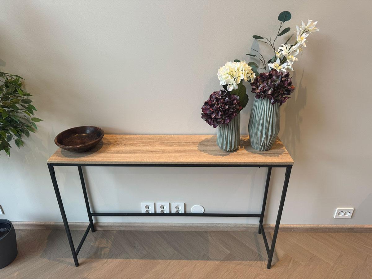 LVT-gulv i lys eikefarge lagt i fiskebensmønster, vist med et smalt konsollbord i tre og sort metall, dekorert med vaser og blomster.