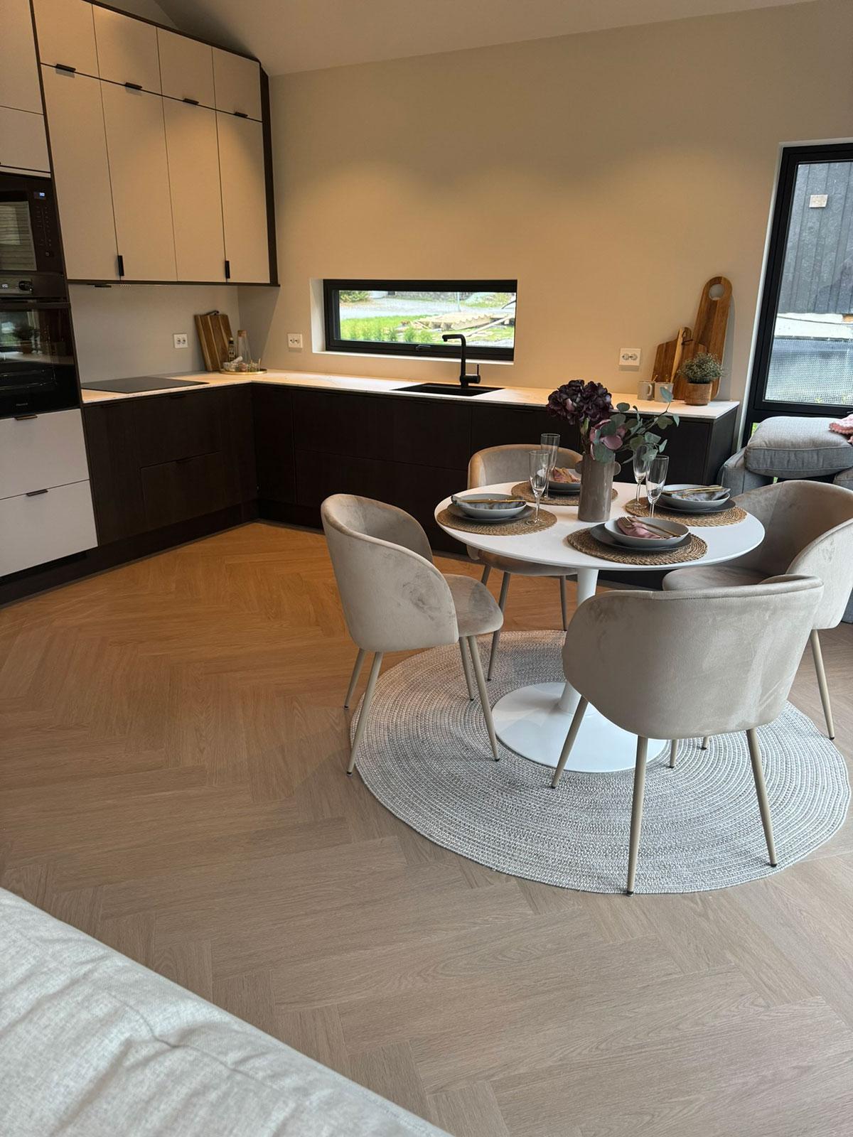 LVT-gulv i lys eikefarge lagt i fiskebensmønster, vist i et moderne kjøkken med mørke fronter og spiseplass med runde former og lyse tekstiler.