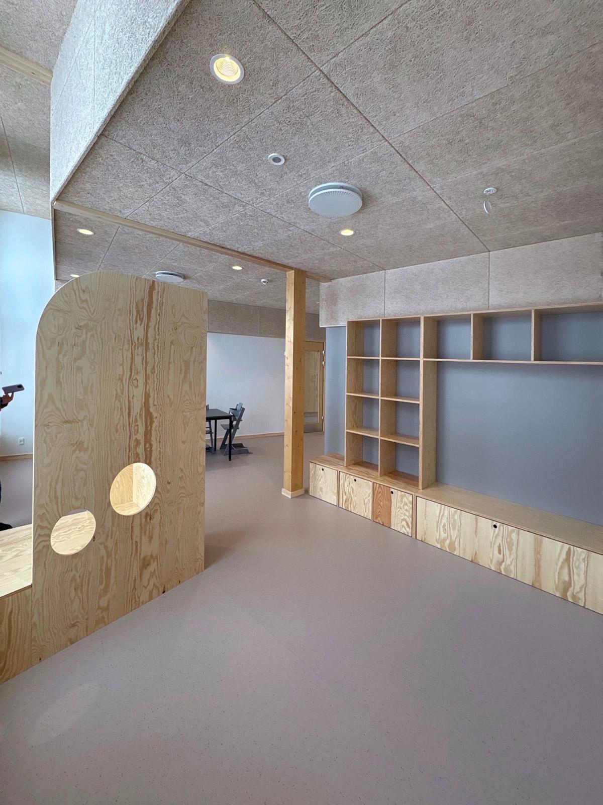 Lyst oppholdsrom med grått gulvbelegg, kryssfinerinnredning, buet lekeskillevegg og åpne trehyller.