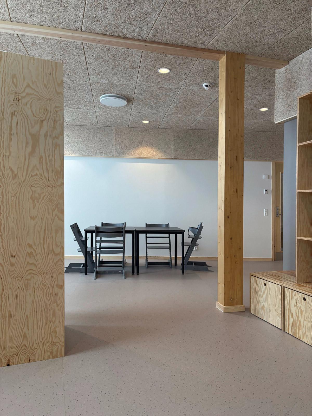 Lyst oppholdsrom med grått gulvbelegg, kryssfinervegger, trehylle og små bord med stoler.