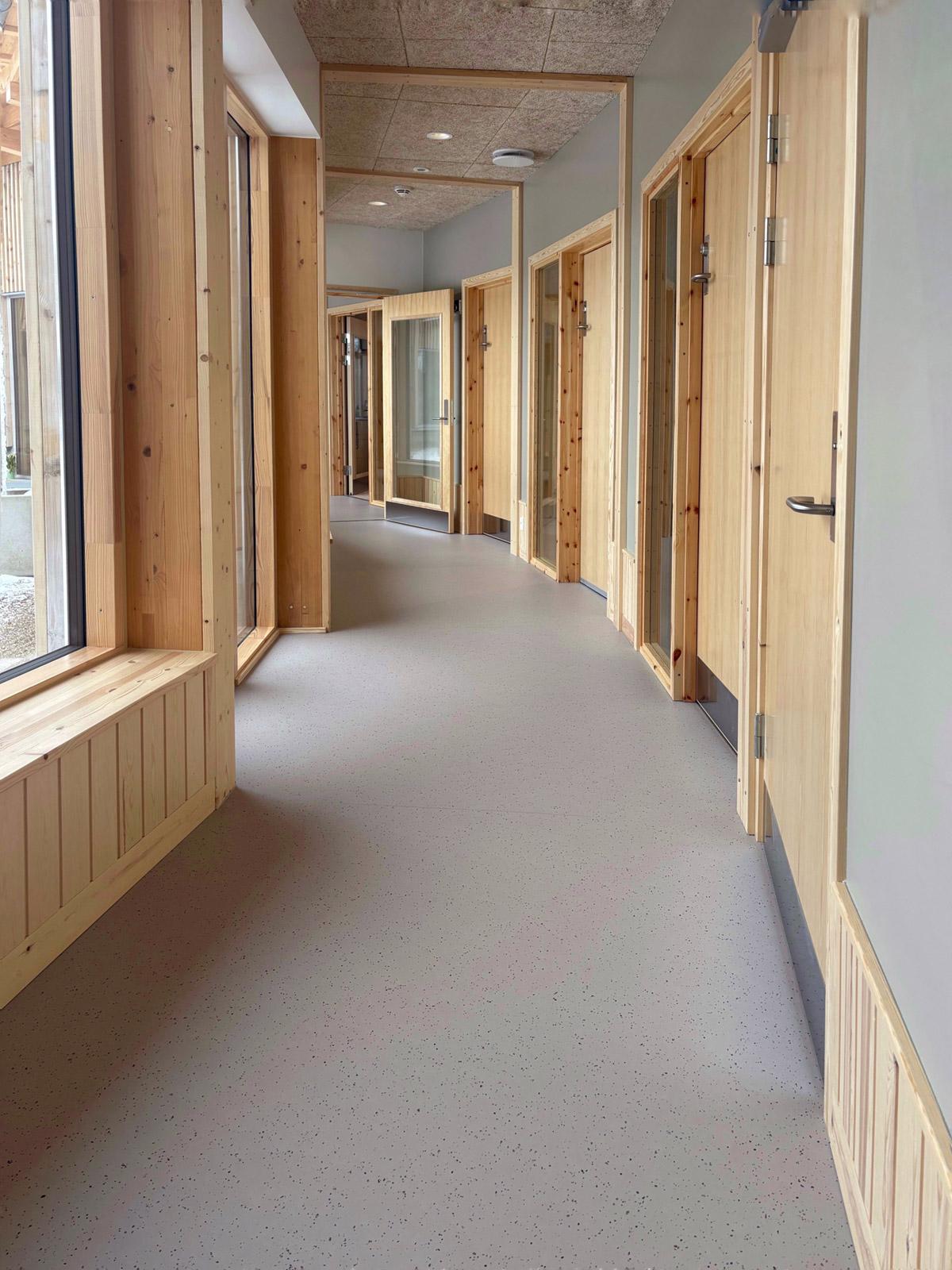 Korridor med grått gulvbelegg, store vinduer og flere rom med dører og karmer i lyst treverk.