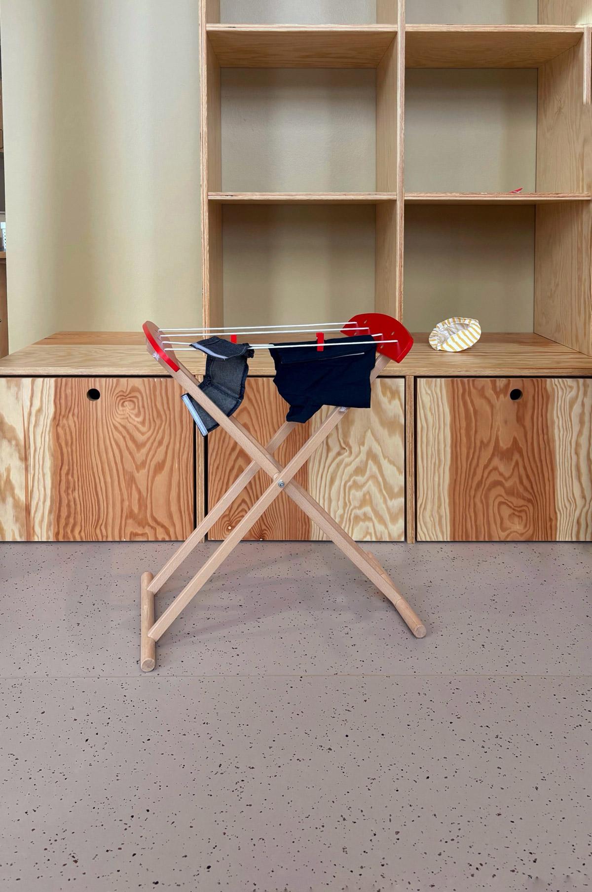 Lekerom med grått prikket gulvbelegg, kryssfinerhyller og lite tørkestativ med barneklær.