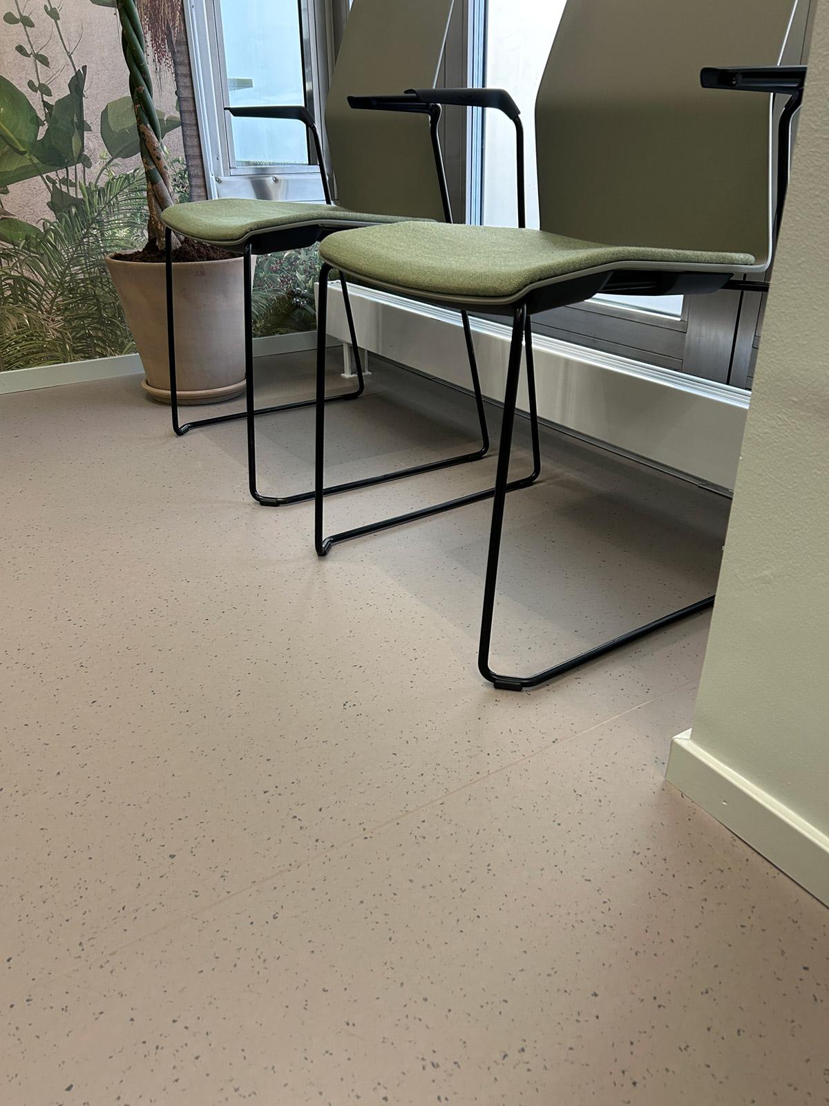 Ventestoler med grønne puter, sort metallramme, plante og lyst prikket gulvbelegg ved store vinduer.