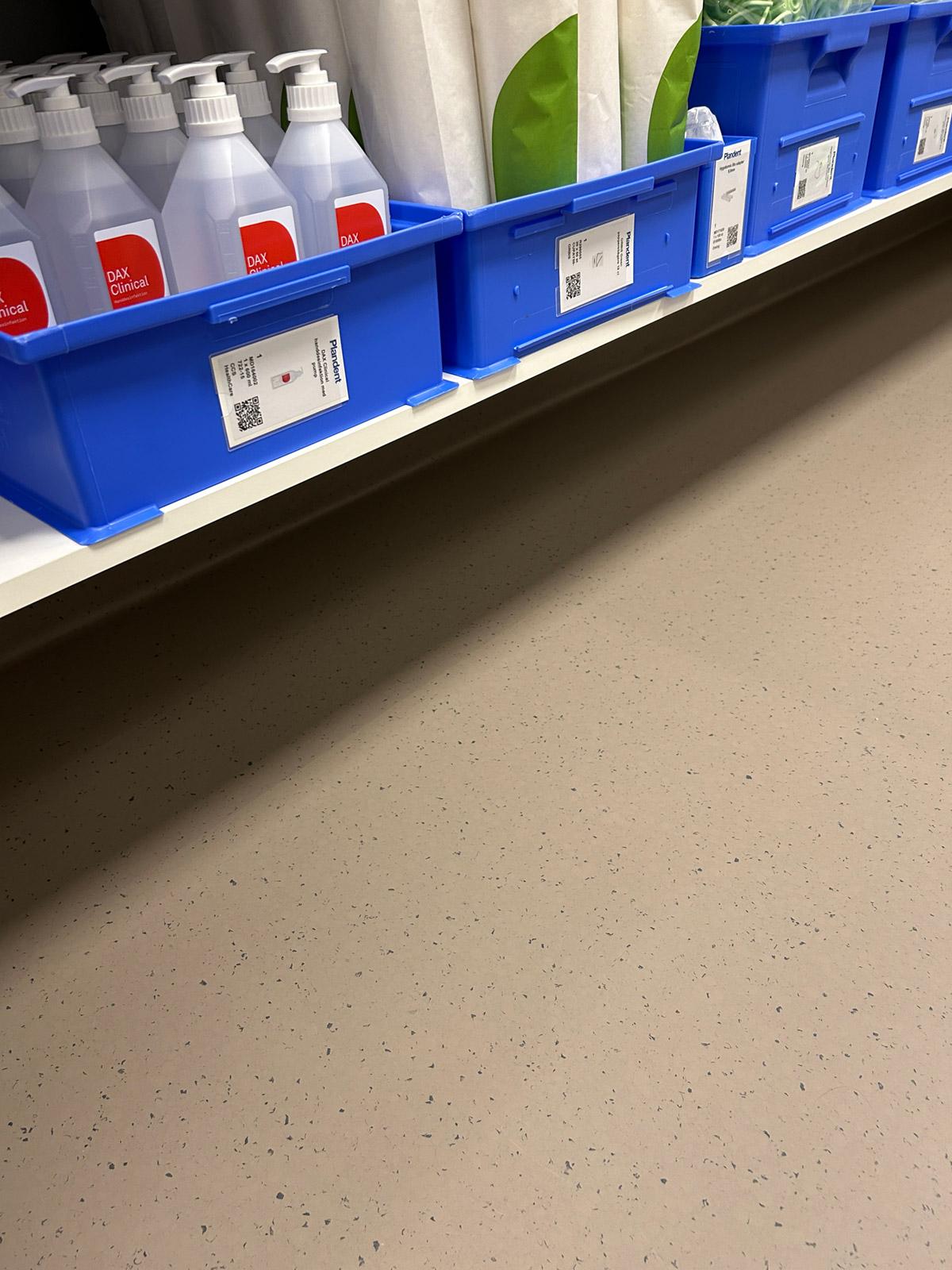 Hylle med blå oppbevaringsbokser, rengjøringsartikler og lyst prikket gulvbelegg.