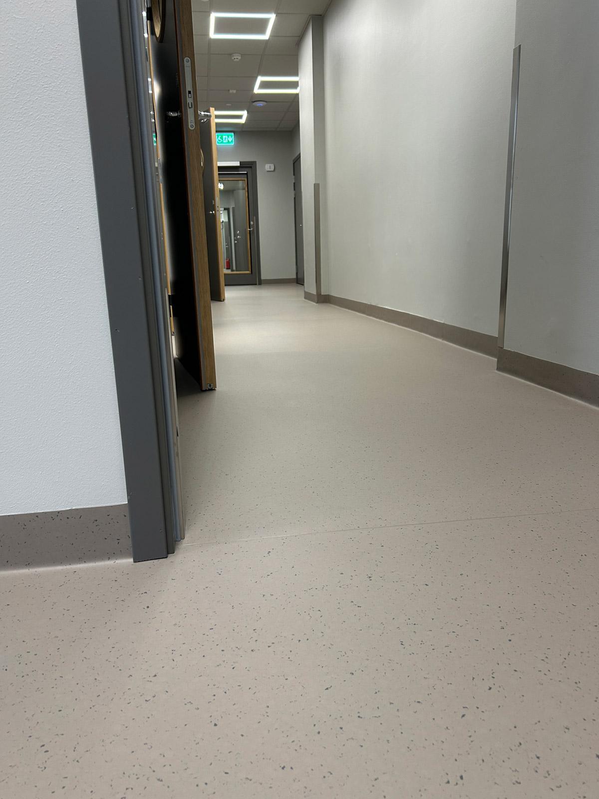Korridor i klinikklokale med lyse vegger, åpne dører og lyst gulv med diskret prikkmønster og oppbrettet sokkel.